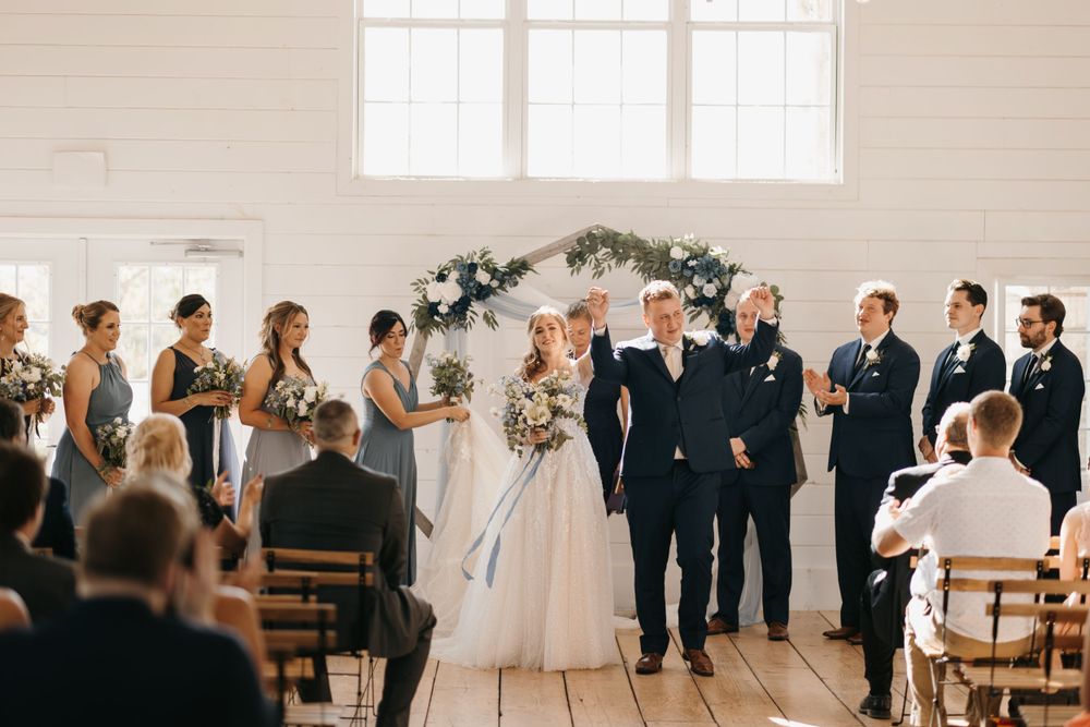 Newlyweds walk back down the aisle as guests applaud at bright white indoor wedding venue.