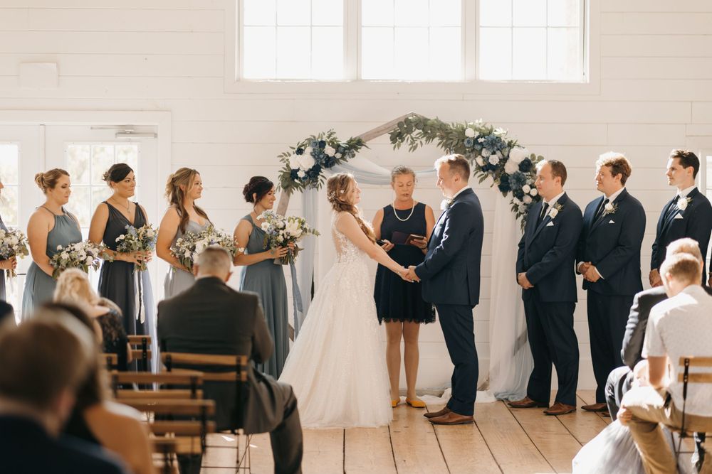 Wedding ceremony with bridesmaids in gray dresses and groomsmen in navy suits at floral arch altar.