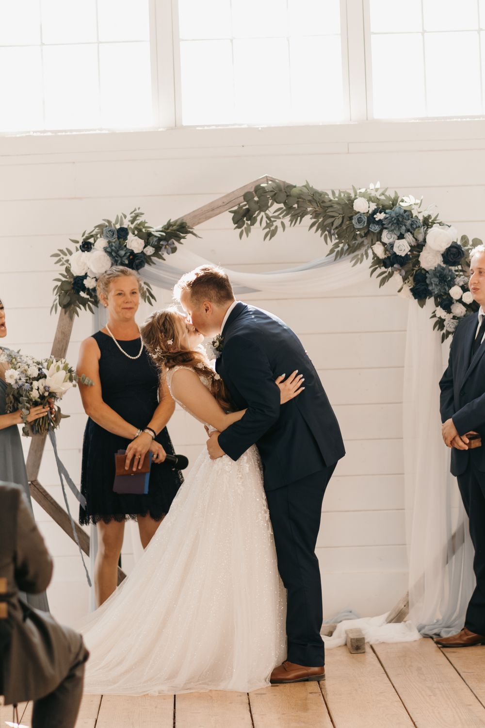 Bride and groom share first kiss at indoor wedding ceremony beneath a greenery and white floral arch.