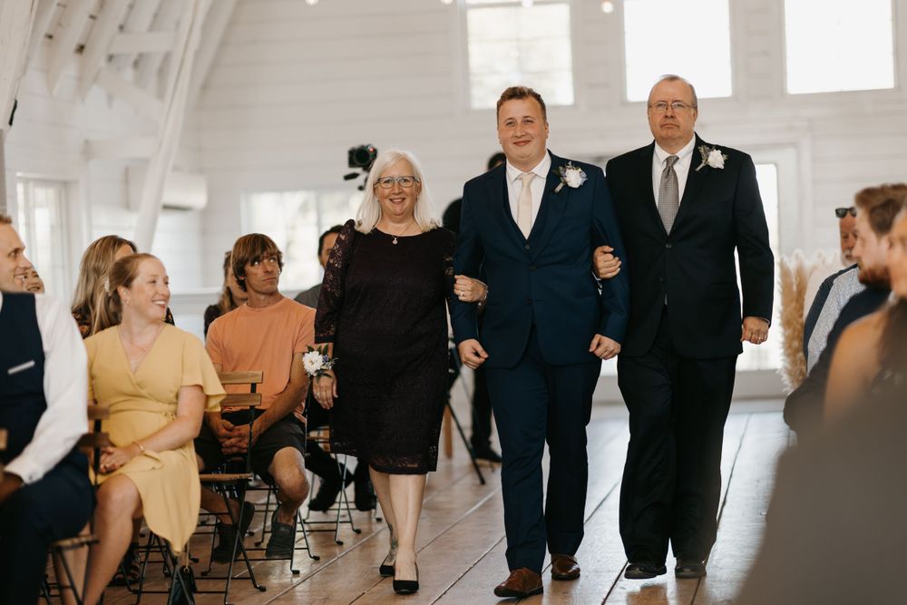 Groom in navy suit is escorted down the aisle by his smiling parents during a wedding ceremony.