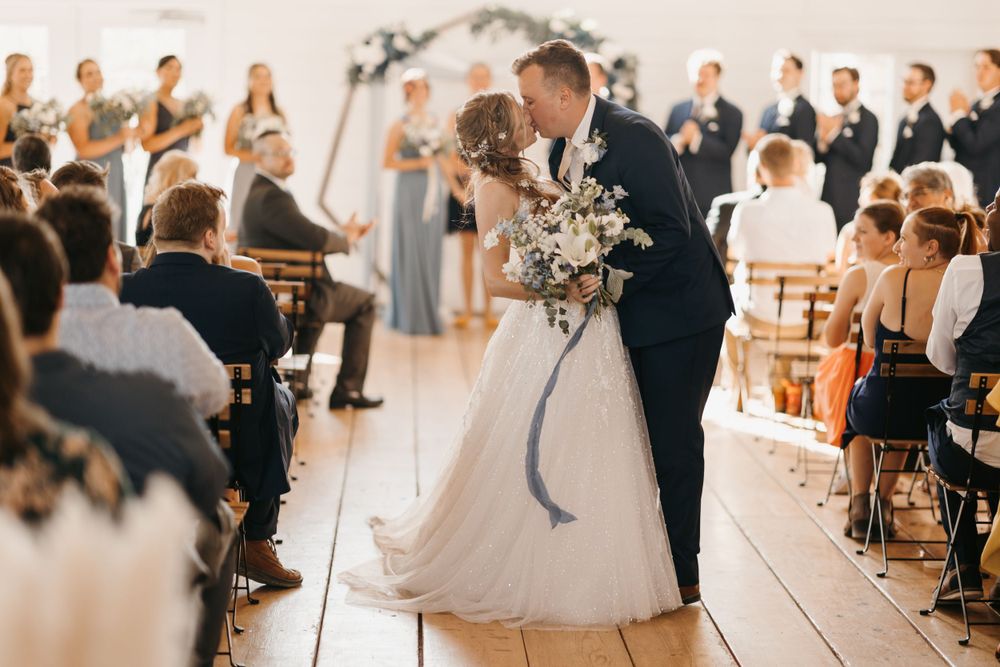 Newlyweds share a romantic kiss in the aisle surrounded by seated wedding guests in white venue.