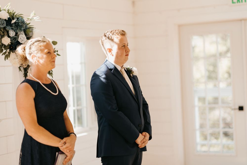 Groom stands at the altar in a navy suit beside a woman in black during an indoor wedding ceremony.