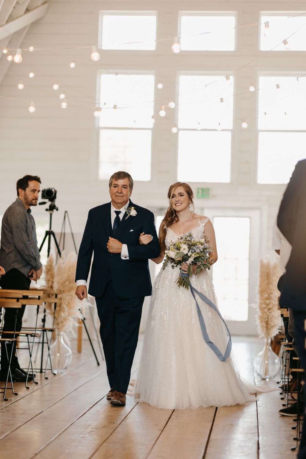 Smiling bride walks arm-in-arm with her father down the aisle in a bright white barn wedding venue.