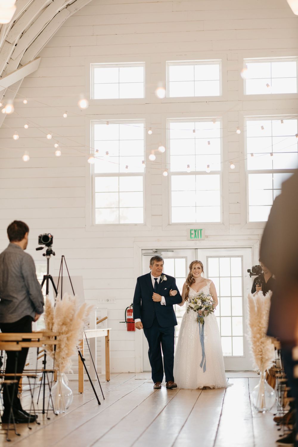 Bride walks down the aisle with her father in a white barn wedding venue decorated with pampas grass.