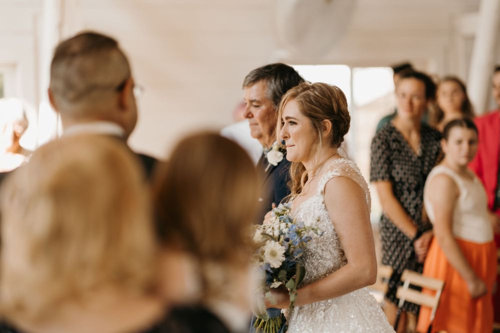 Bride holding floral bouquet walks down the aisle with her father past standing wedding guests.