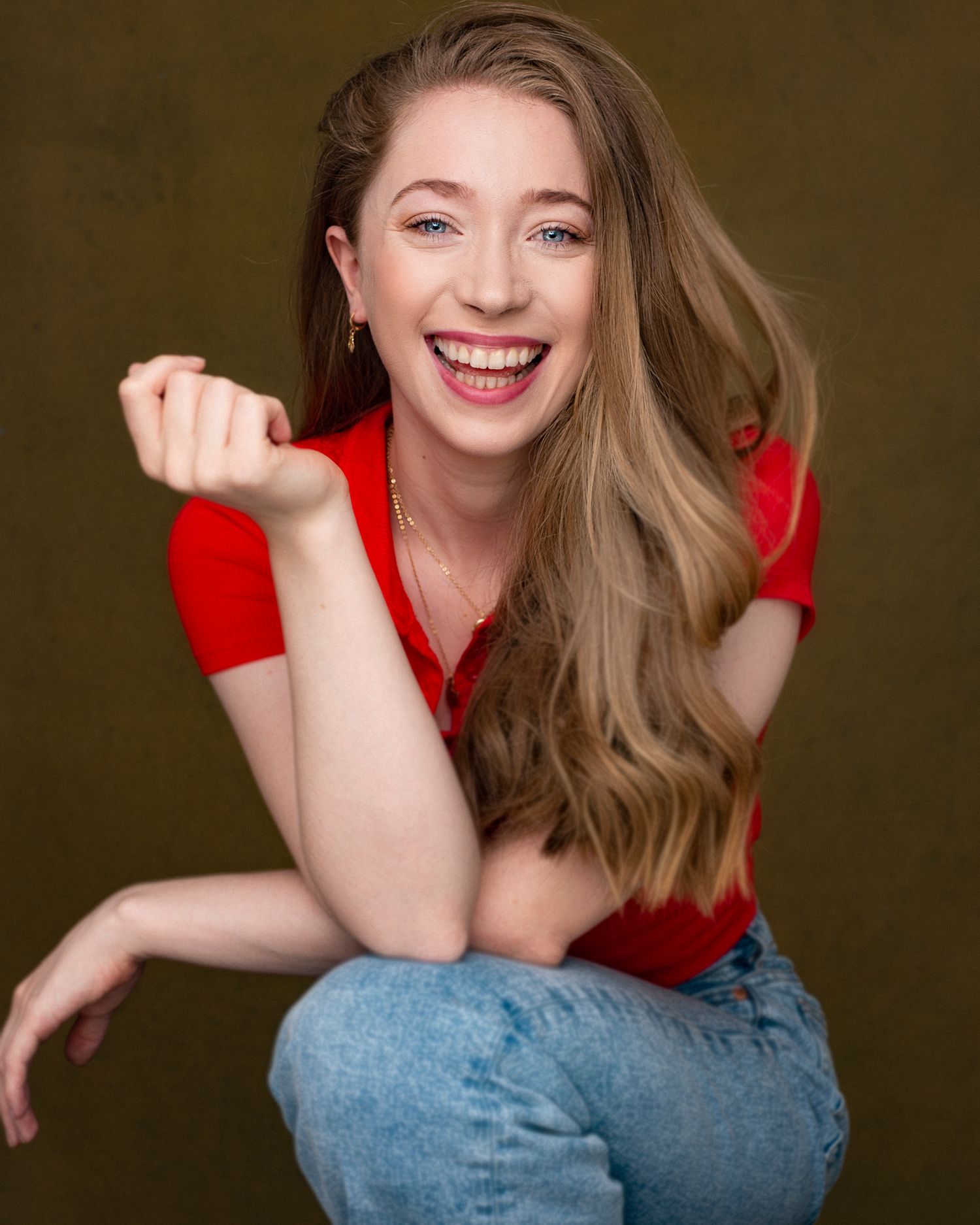 Studio portrait of someone in a red top and jeans expressing joy against a brown backdrop.