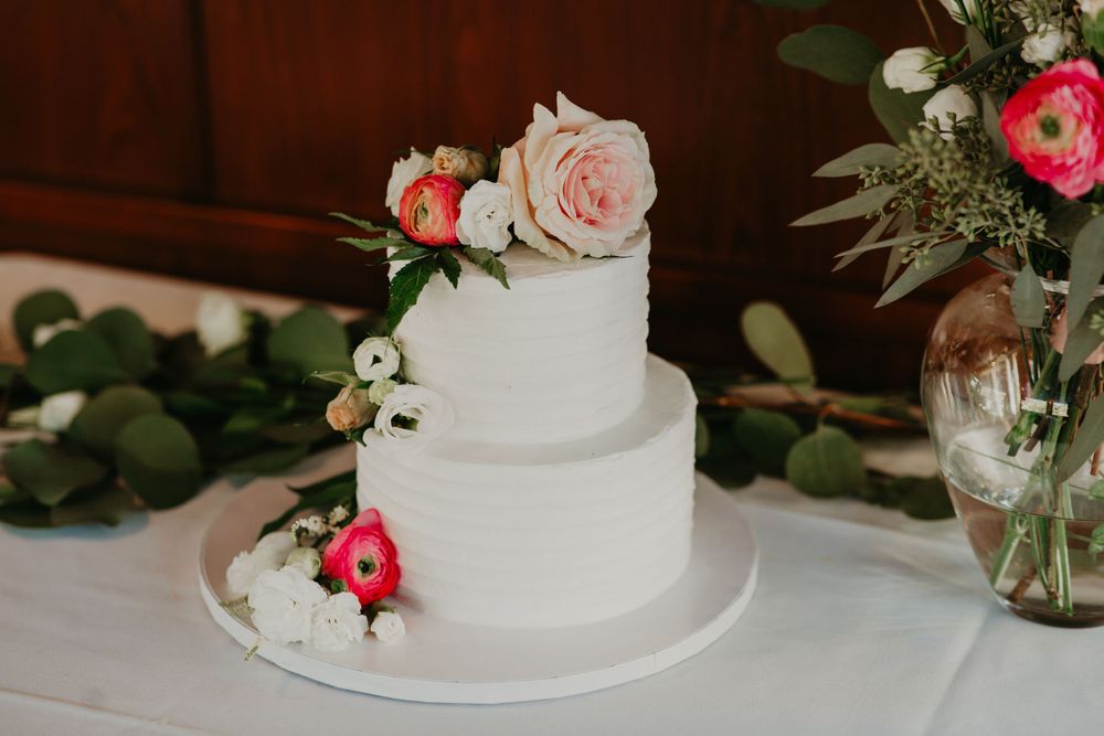 Two-tier white wedding cake adorned with pink and red flowers on a table with greenery decor.