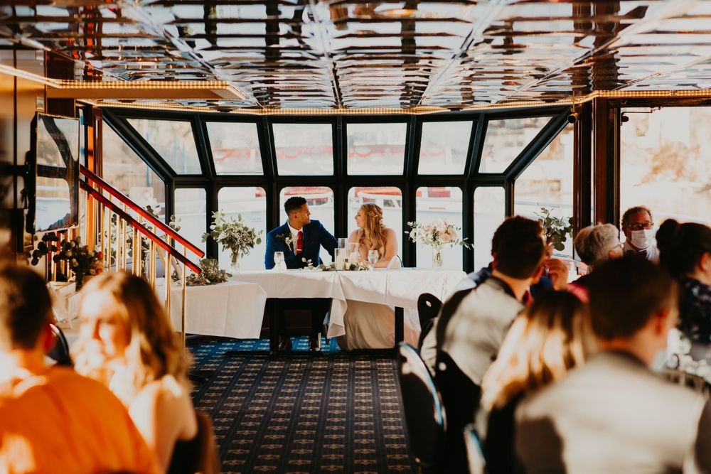 Wedding reception dinner on a boat with bride and groom seated at head table surrounded by guests.