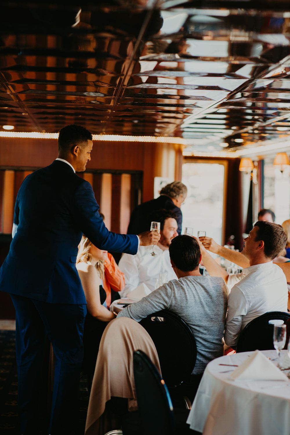 Groom in blue suit toasting champagne with seated wedding guests at reception dinner.