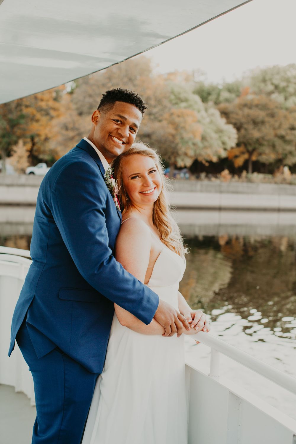 Happy bride and groom hold hands on a boat deck surrounded by fall foliage.