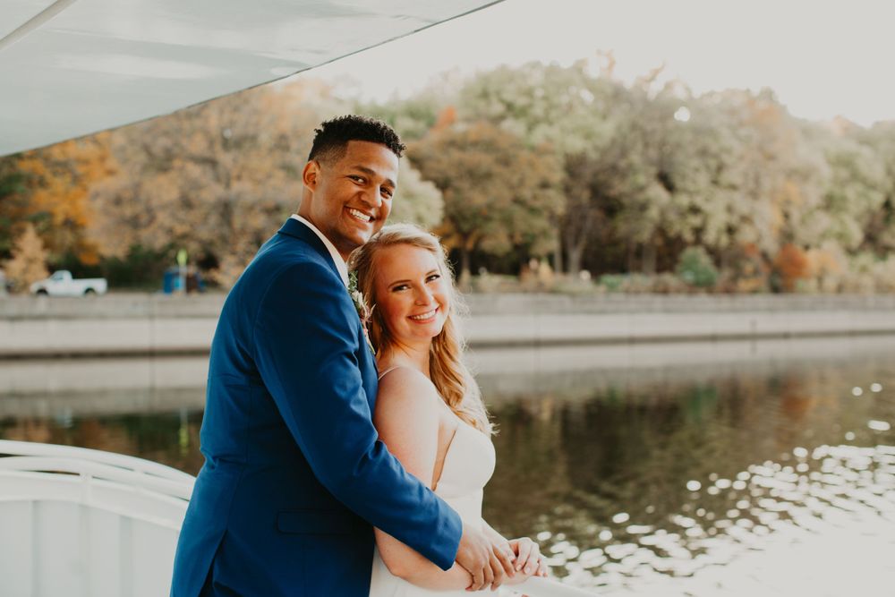 Smiling newlyweds pose together on a boat deck with autumn trees reflected in the water.