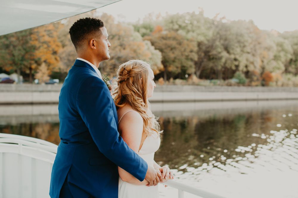 Bride and groom hold hands while gazing at a river surrounded by colorful fall foliage.