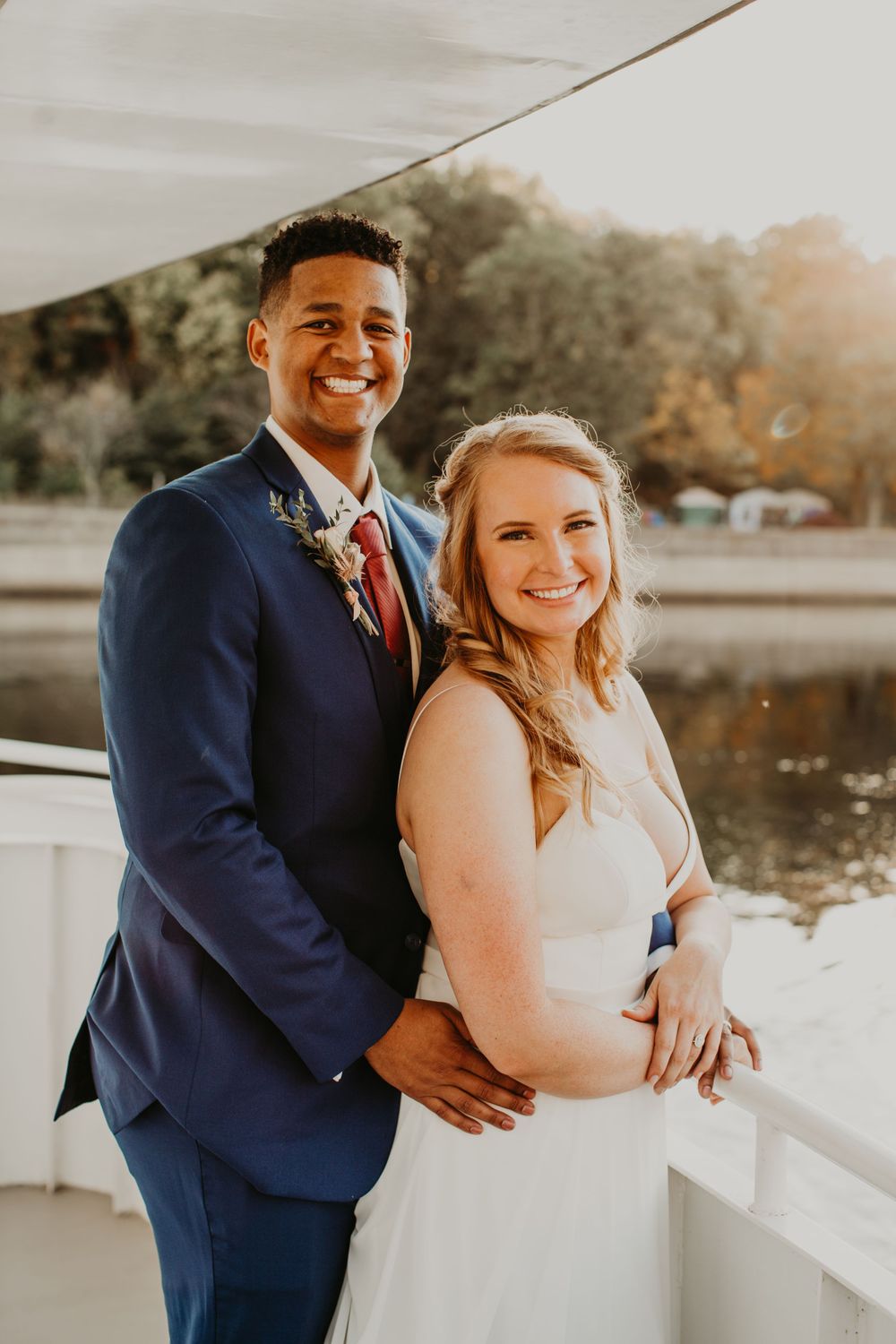 Smiling interracial newlyweds pose on a sunlit boat deck, groom in navy suit with red tie, bride in white wedding gown.