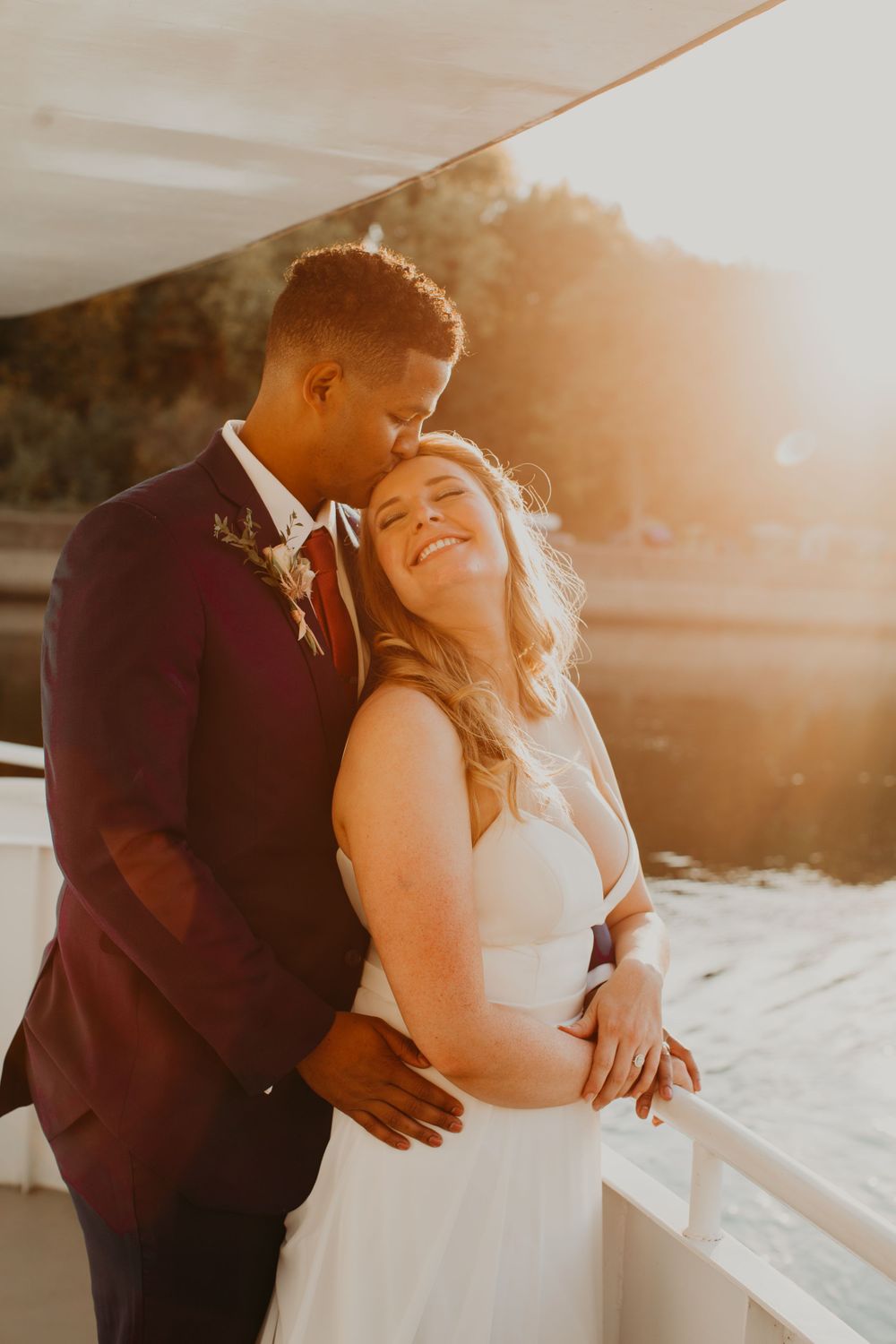 Groom in burgundy suit tenderly kisses bride on forehead in golden sunset light on a boat deck.