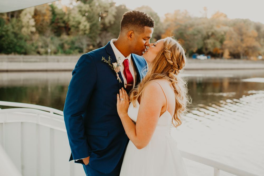 Interracial bride and groom share a romantic kiss on a white boat deck with river backdrop.