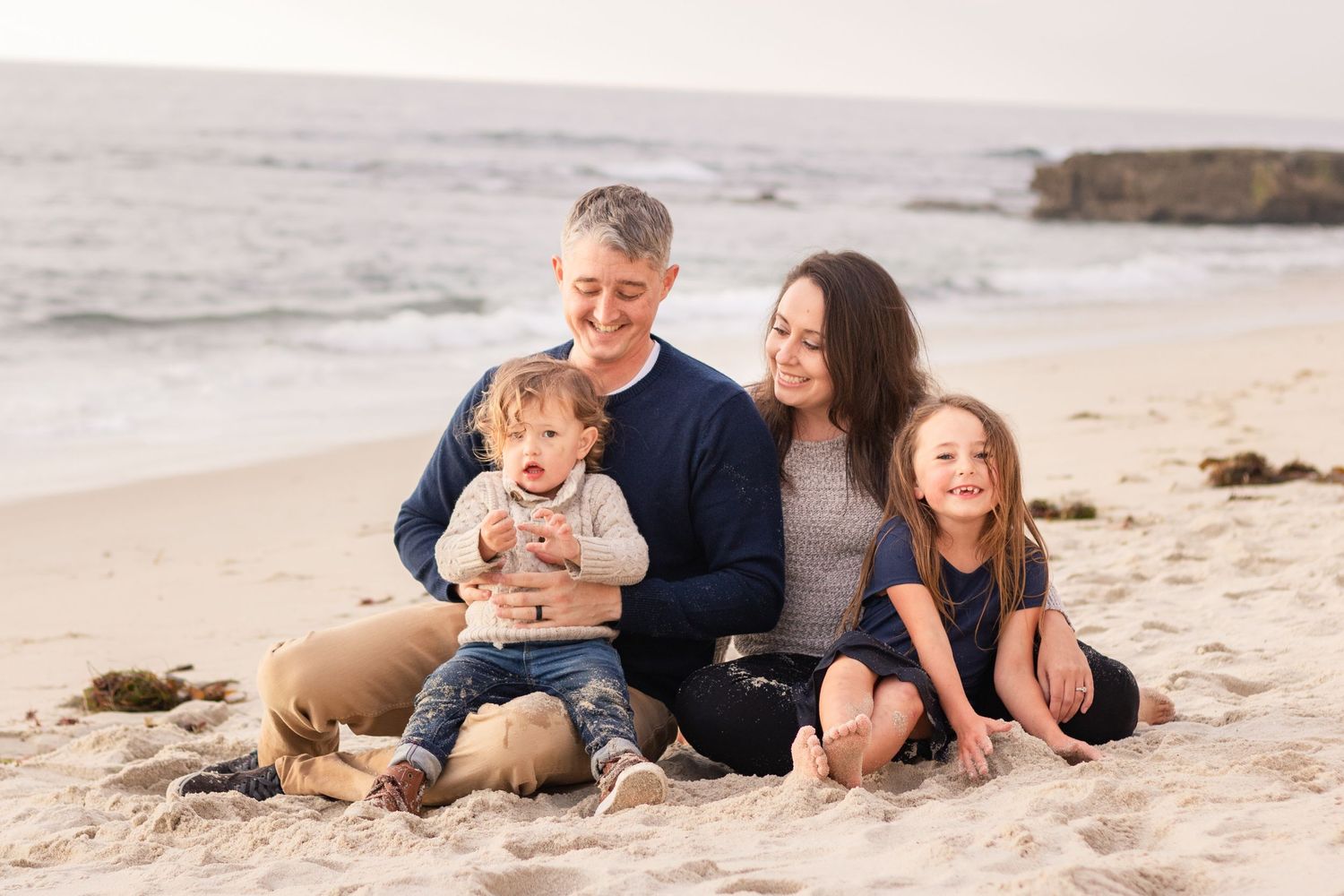 Family of four sits together on sandy beach at sunset near rocky coastline.