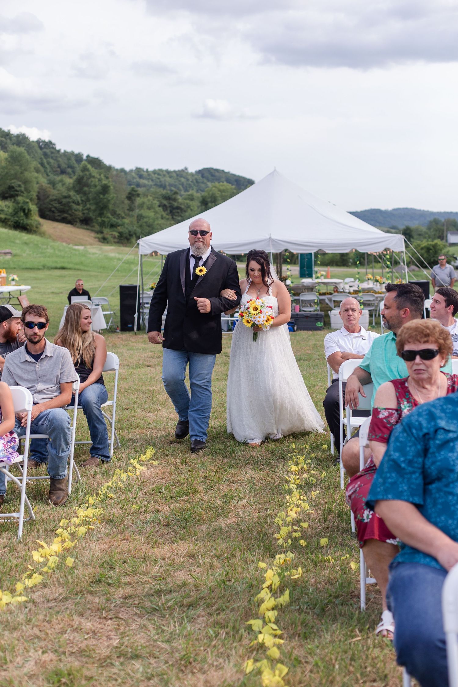 Outdoor wedding ceremony under a white tent with guests seated in chairs decorated with sunflowers.