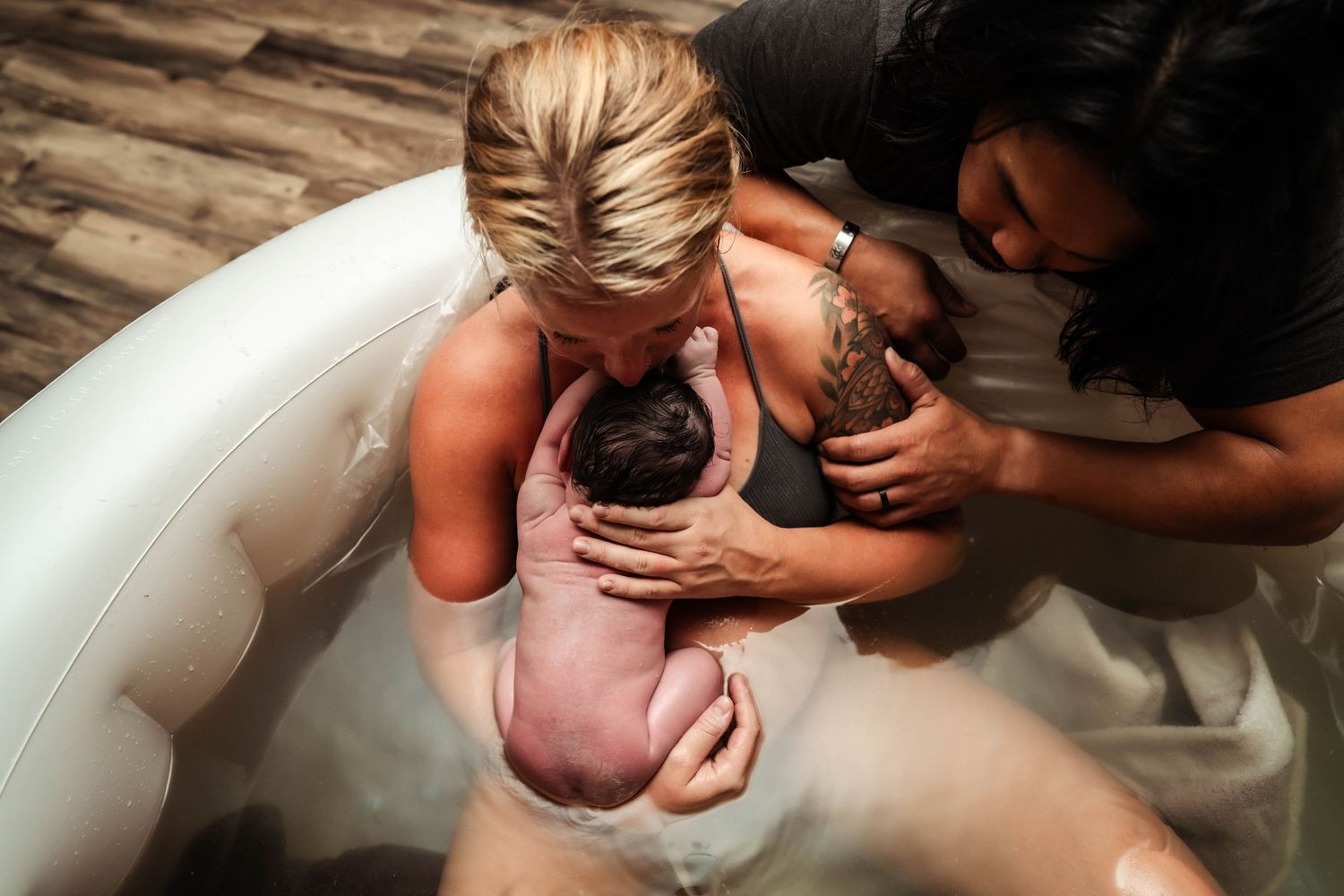 Woman in birth pool holding newborn baby. Her husband is looking over her shoulder with hand on her arm.