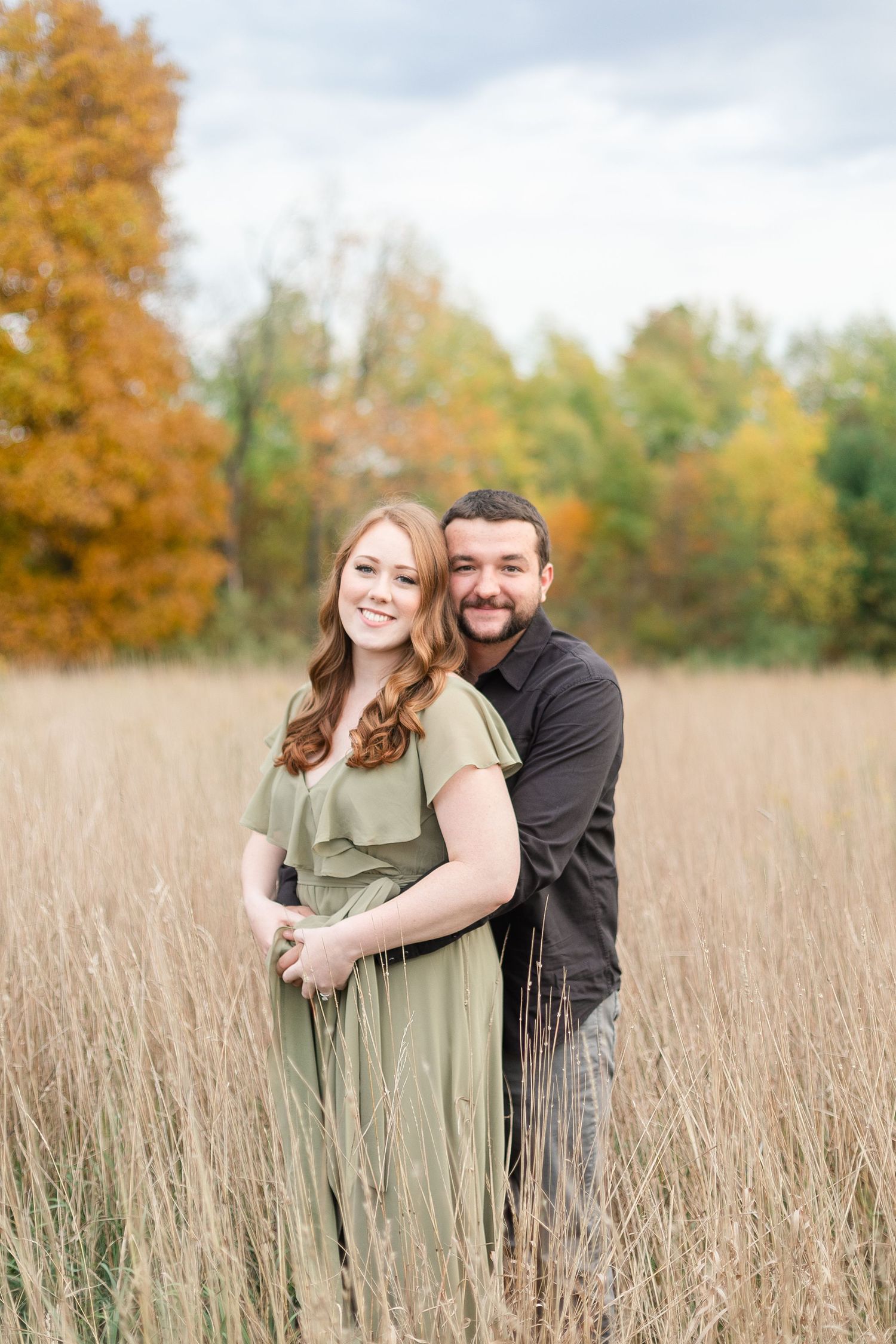 A couple embraces in a field of tall grass with autumn foliage in the background.