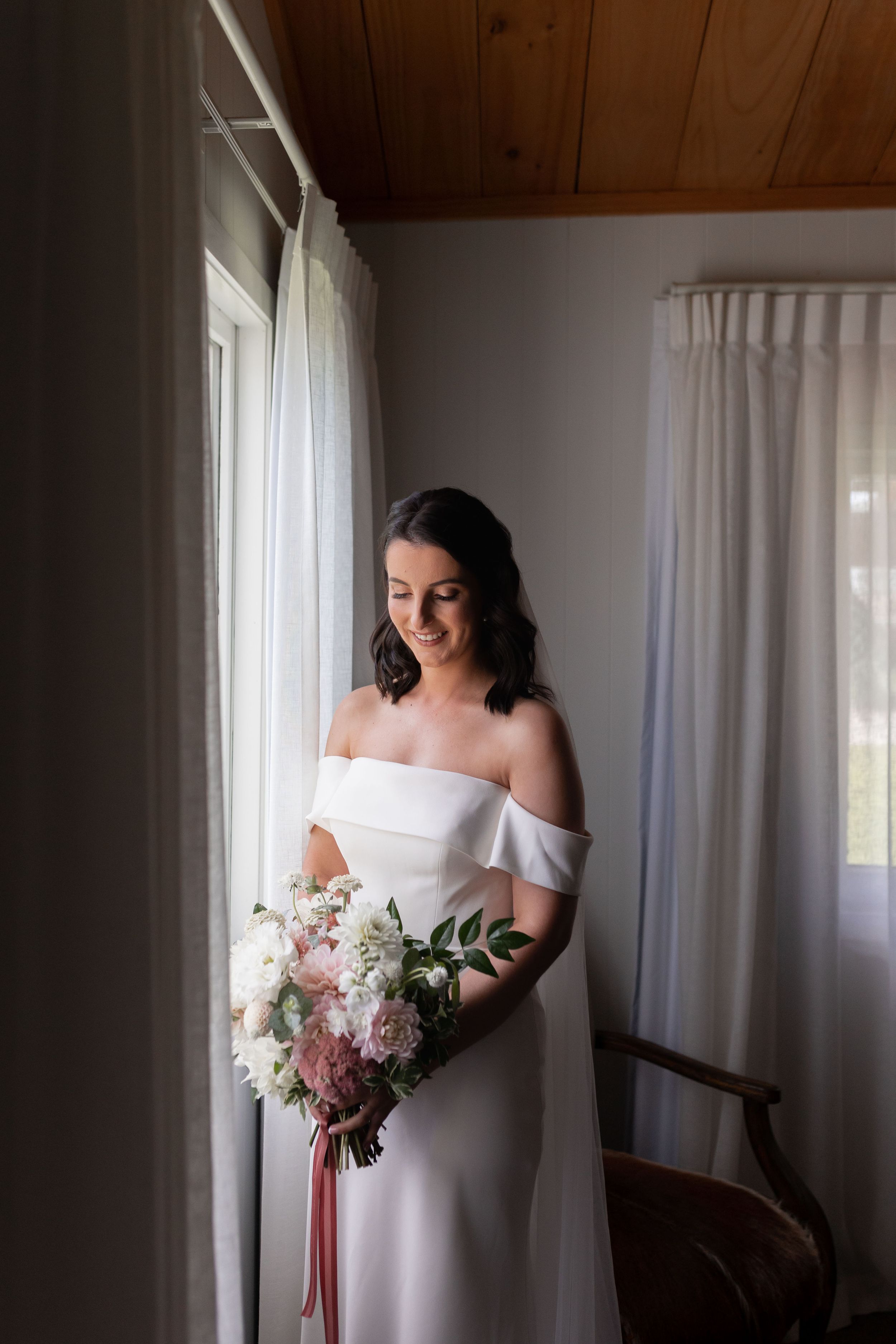 A bride in an off-shoulder white dress holds a bouquet by a window.