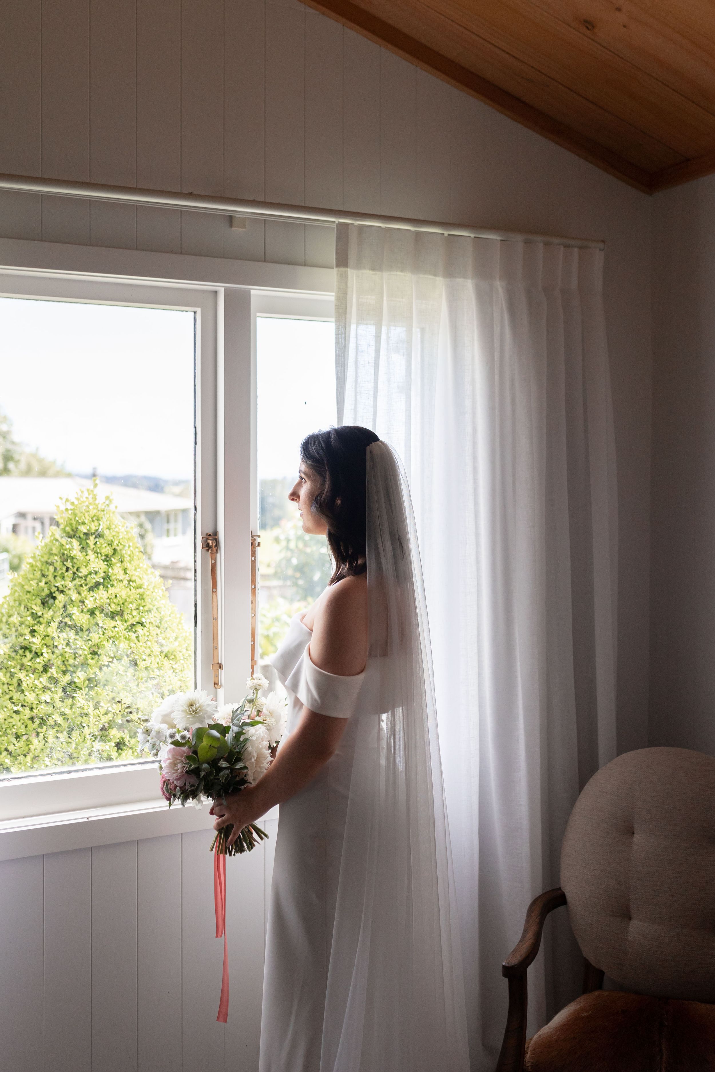A silhouetted figure in a wedding dress stands by a bright window holding flowers.