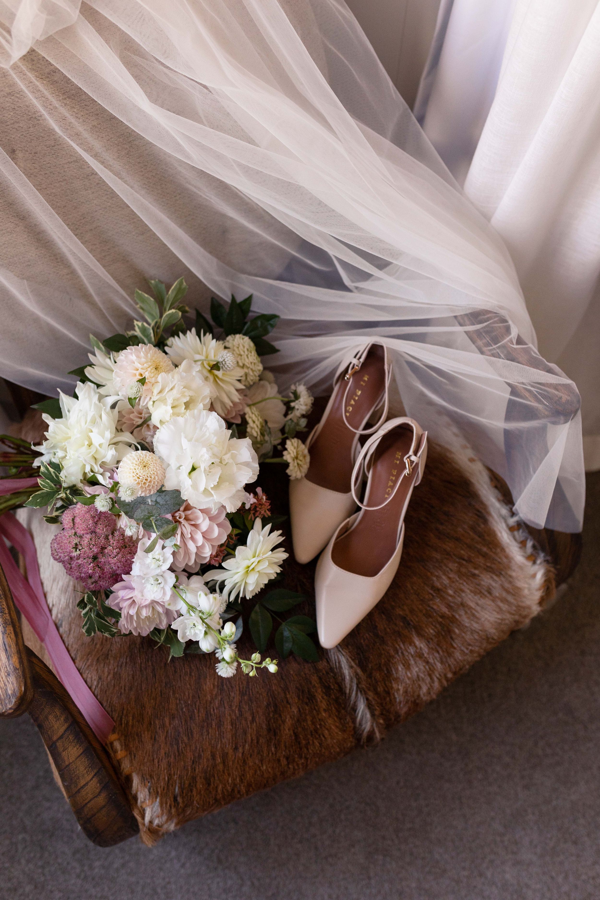 Wedding details with bridal bouquet, veil, and nude heels on rustic wooden surface.