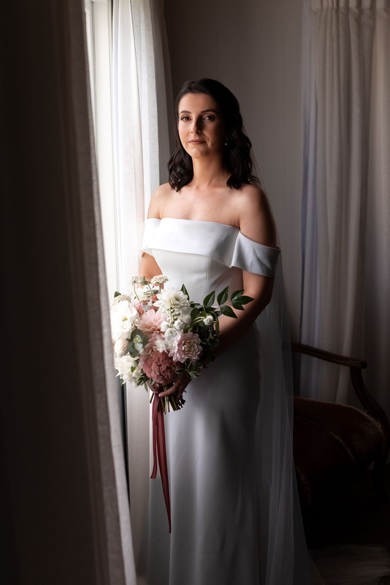 Elegant off-shoulder wedding dress with soft pink and white bouquet in window light.