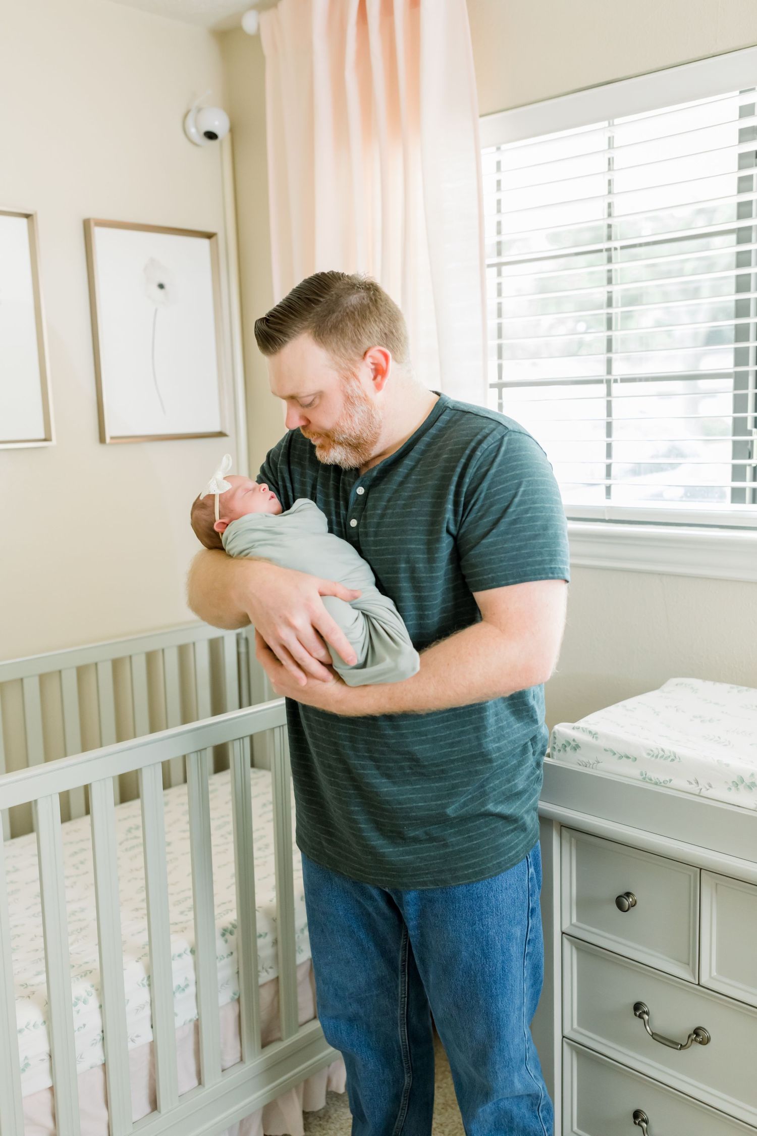 A person in a green shirt tenderly holds a newborn baby wrapped in gray near a nursery crib.