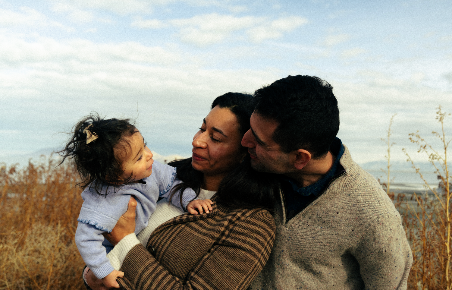 A loving family moment shared outdoors against a backdrop of tall grass and blue sky.