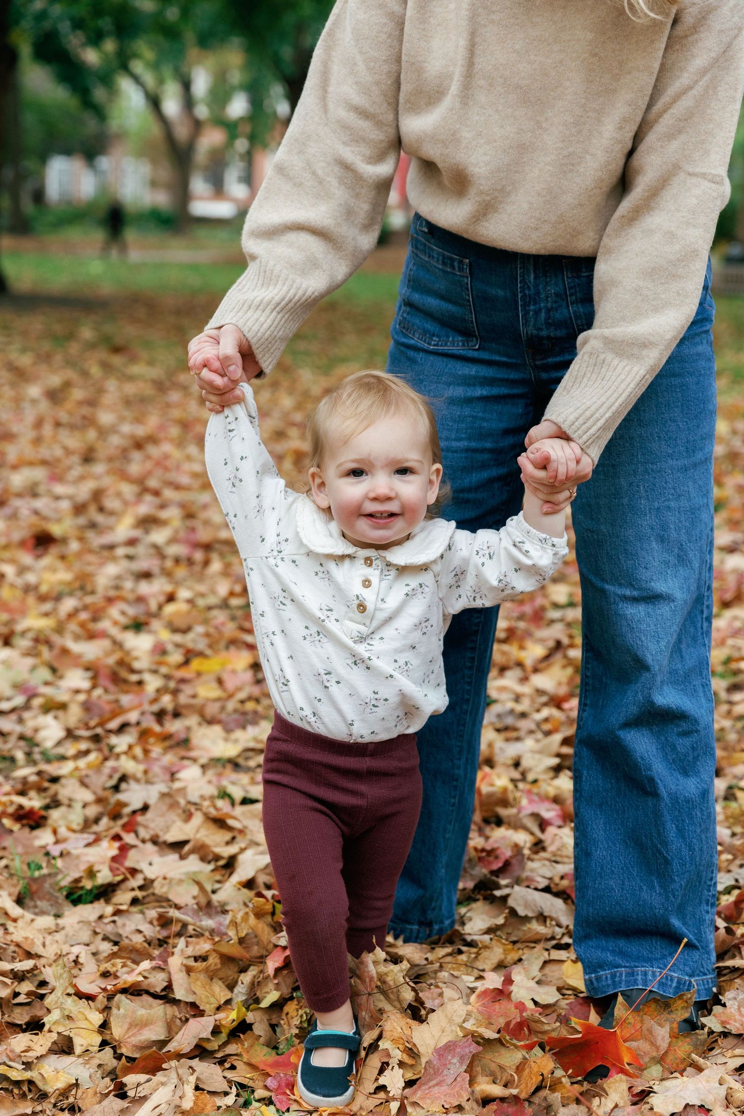 Child learning to walk holds hands with adult through autumn leaves.