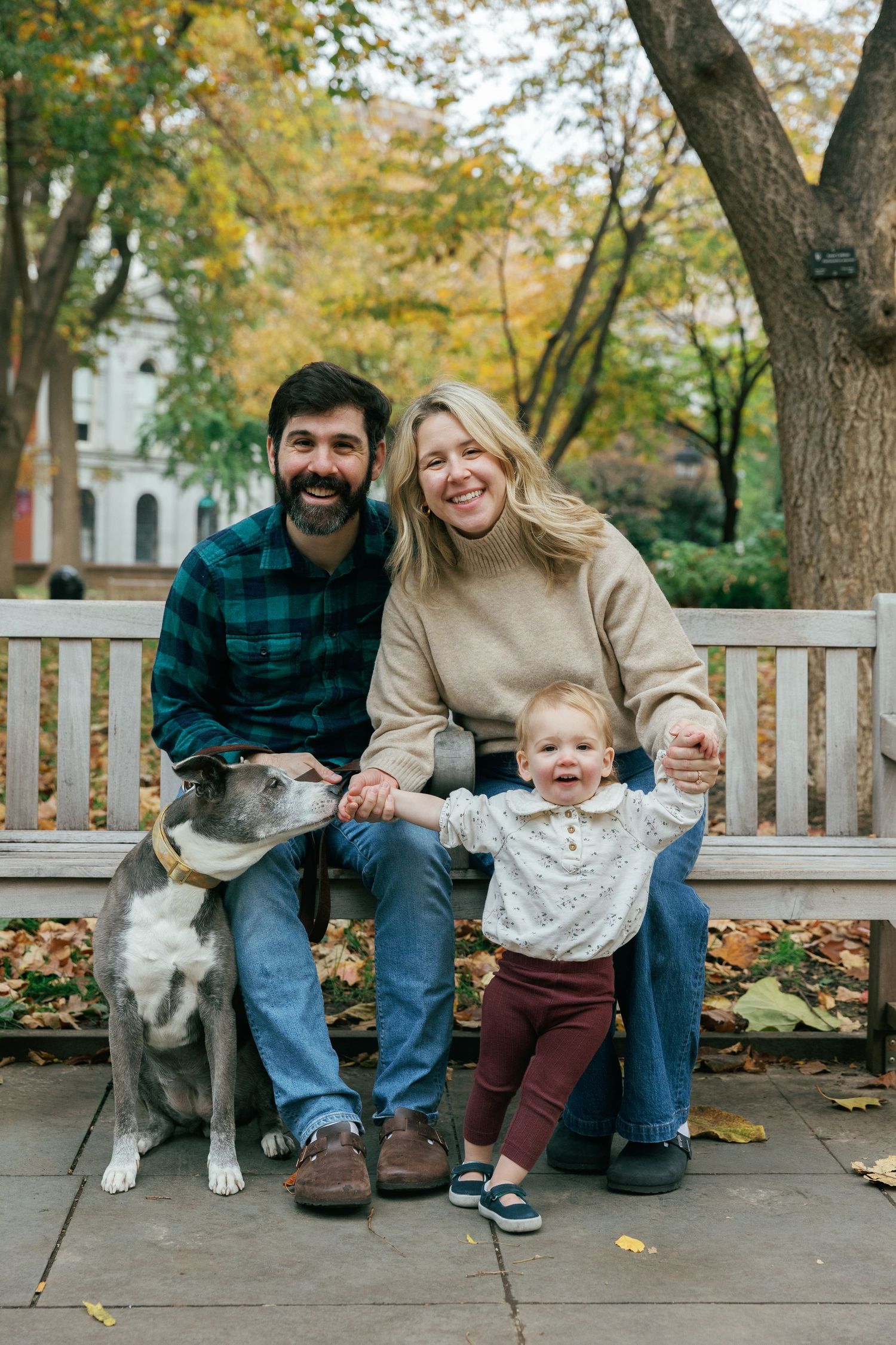 Family and their dog sitting together on park bench surrounded by fall foliage.