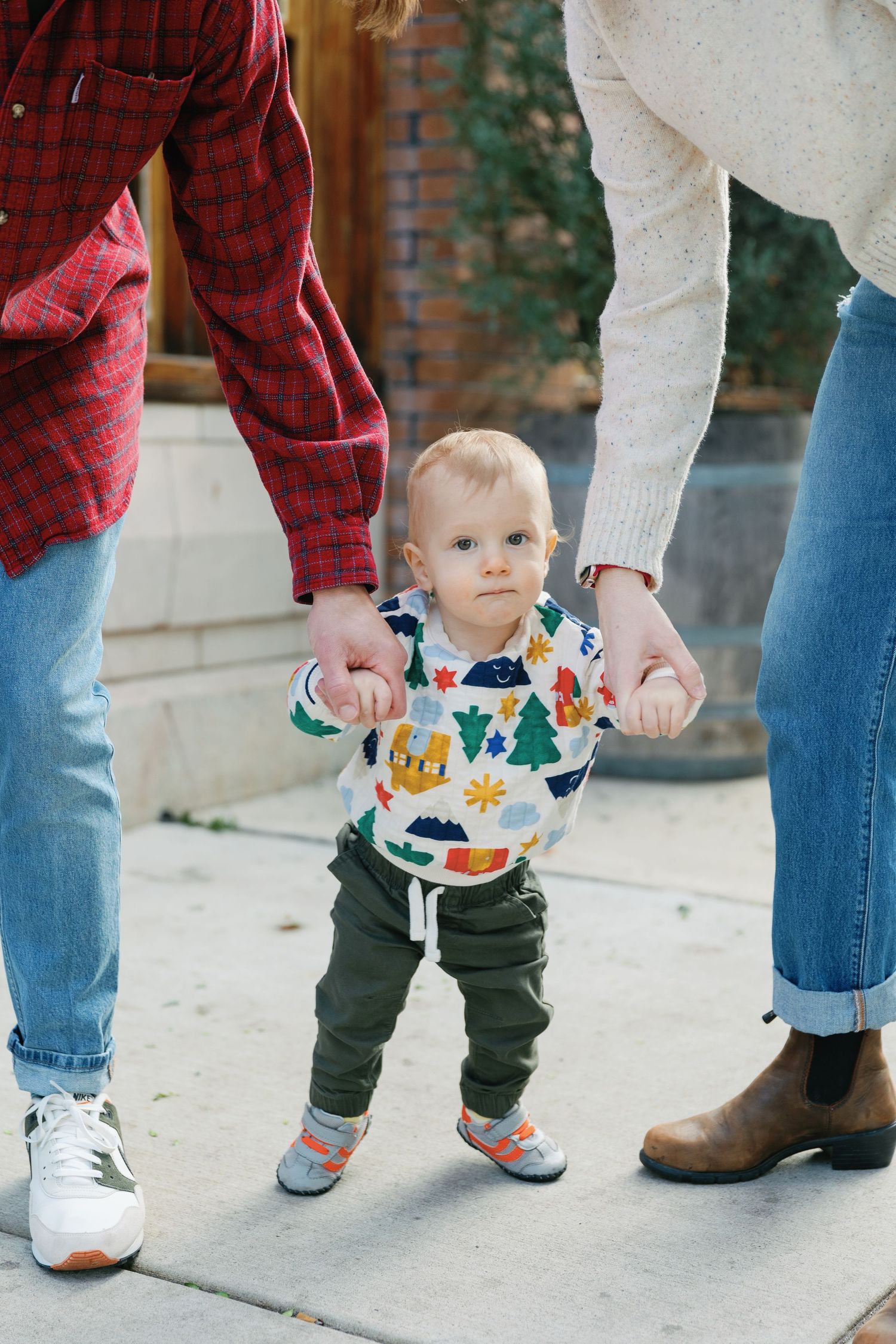 Toddler taking first steps while holding parents' hands on sidewalk.