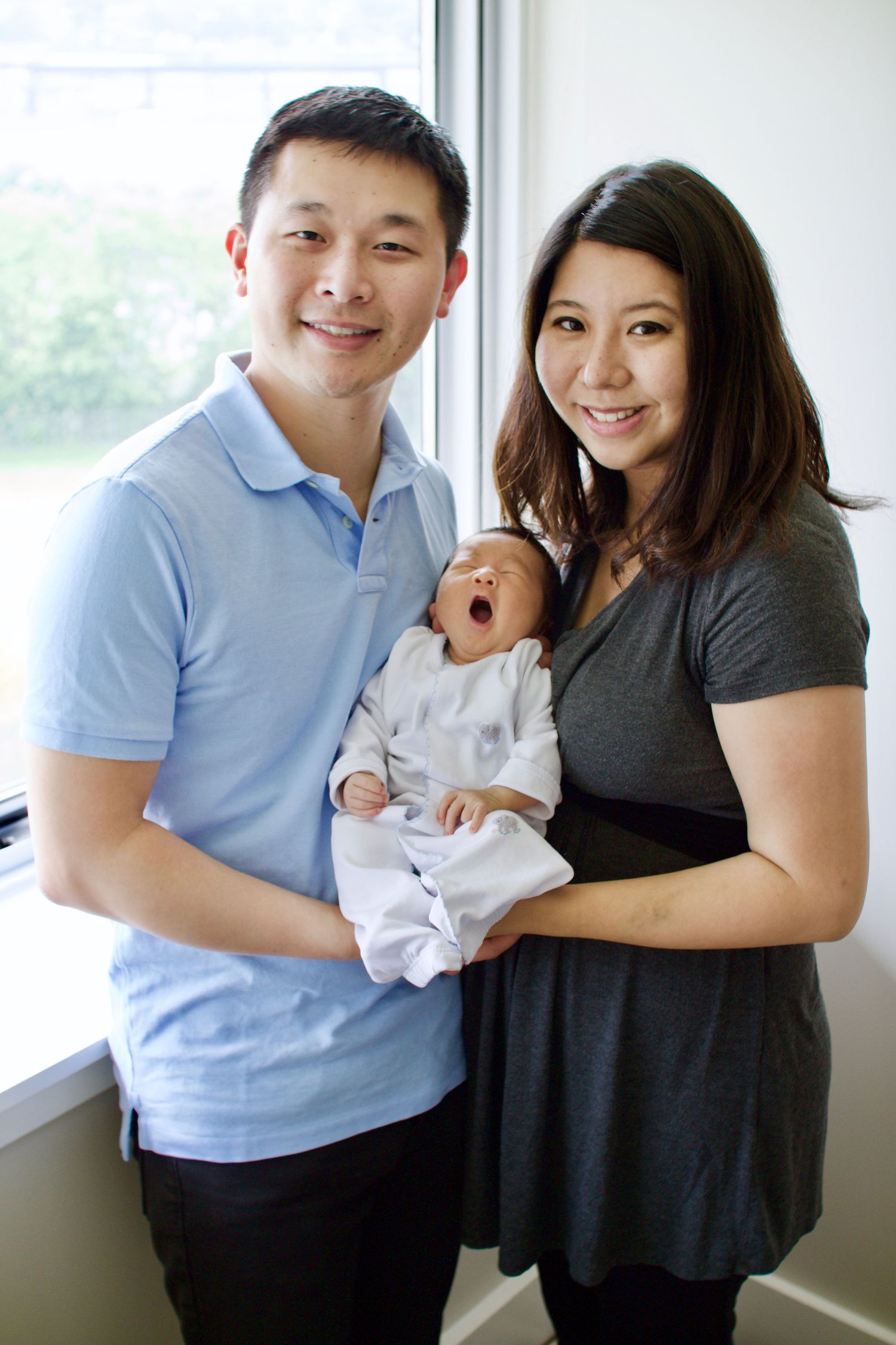 A young couple in casual attire holds their newborn baby near a bright window.