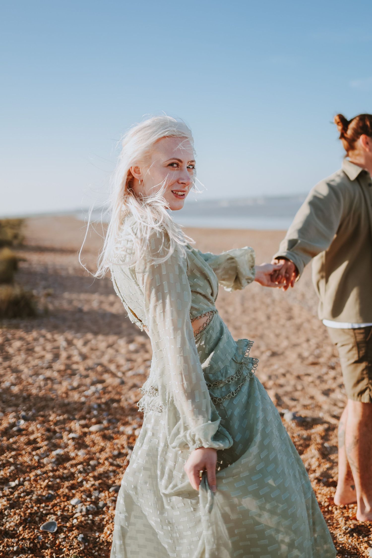 A romantic couple in vintage attire walks along a rocky beach during sunset.