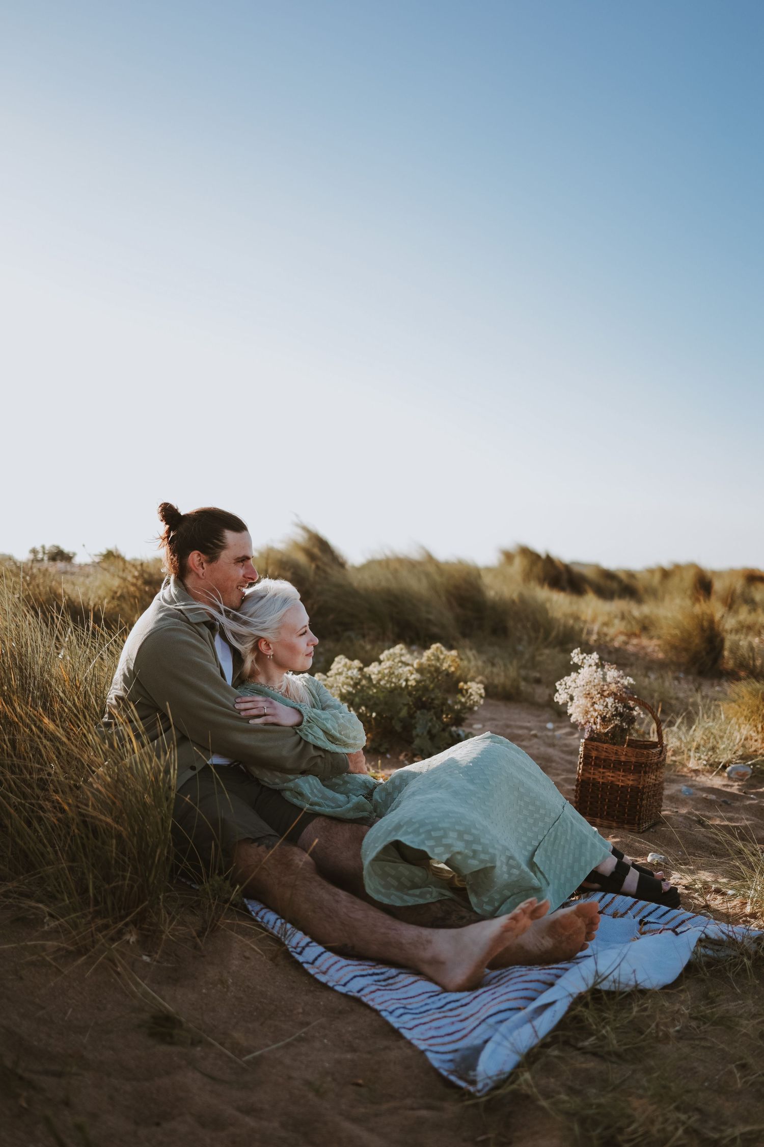 Two people share a picnic blanket among coastal dunes during a sunny afternoon.