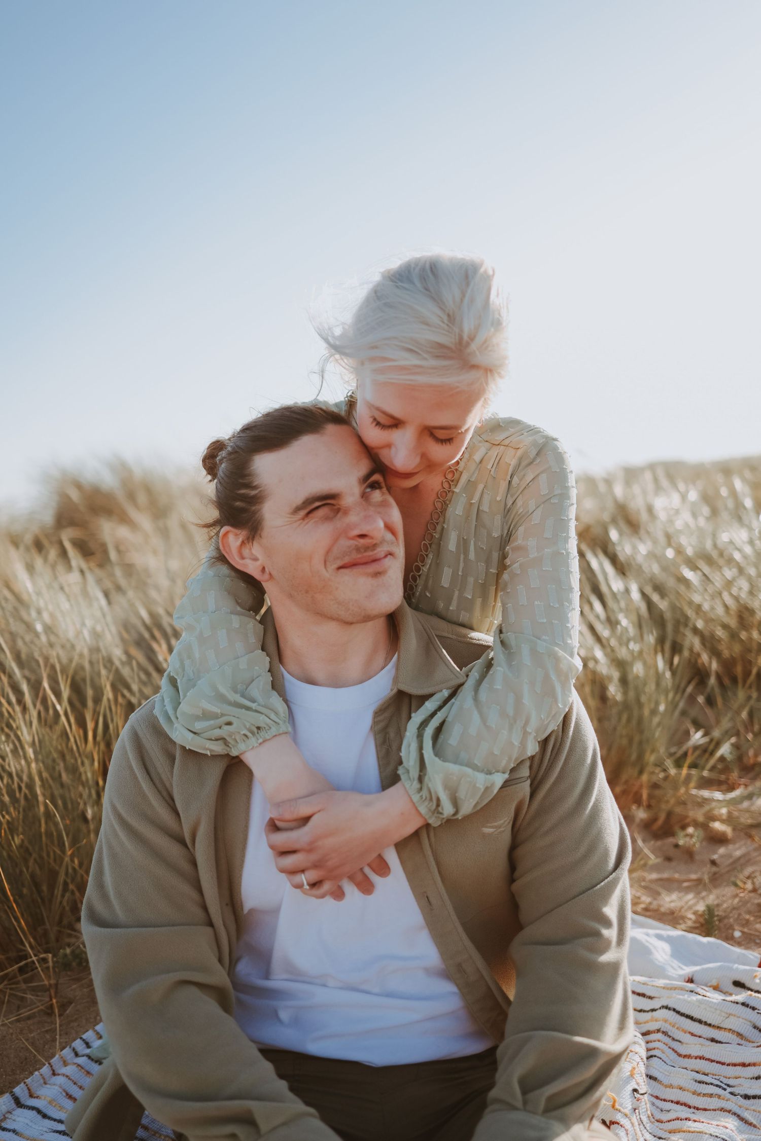 A couple shares a warm embrace while sitting in coastal grassland during golden hour.