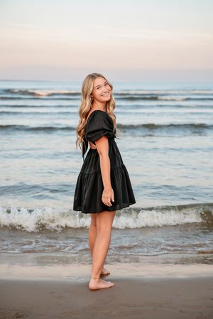 Woman in black dress stands barefoot on beach at sunset with waves in background.