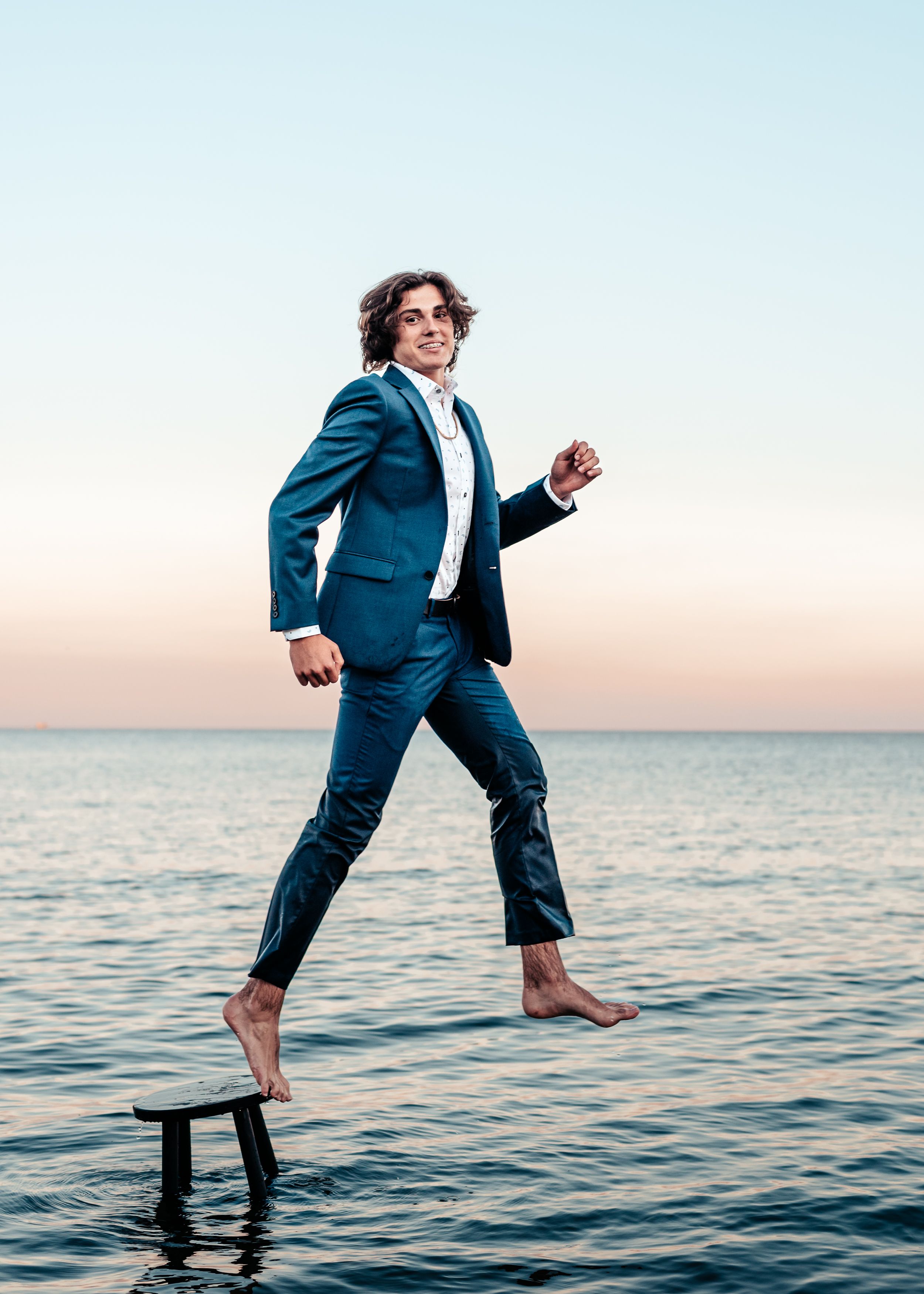 Person in a blue suit leaps dramatically over ocean waters at sunset.