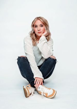 Person in white knit sweater and jeans sits cross-legged against white background wearing tan sneakers.