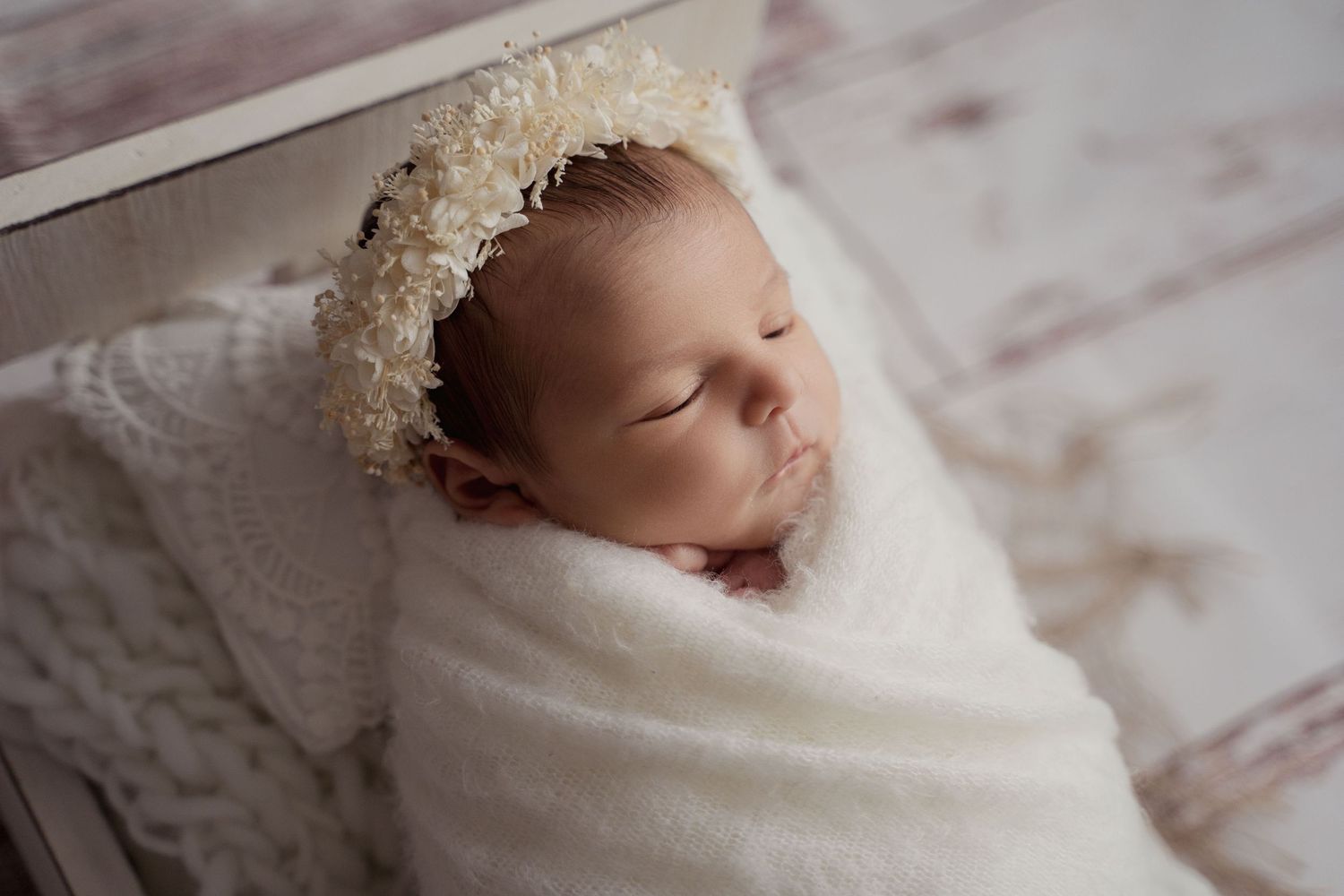 Peaceful newborn baby sleeping wrapped in white swaddle with delicate floral headband.