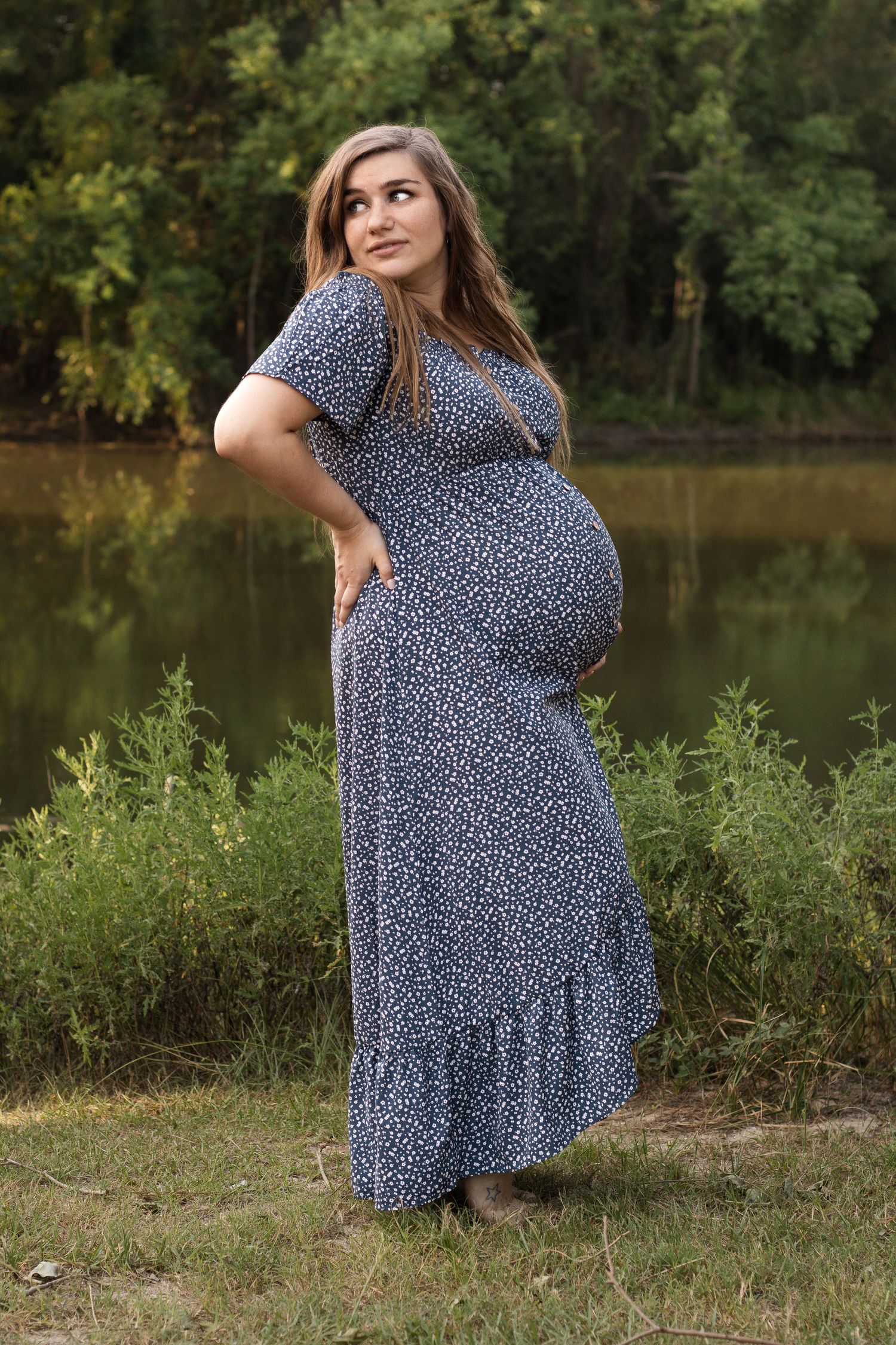 Pregnant woman in floral dress poses by a lake during maternity photoshoot.