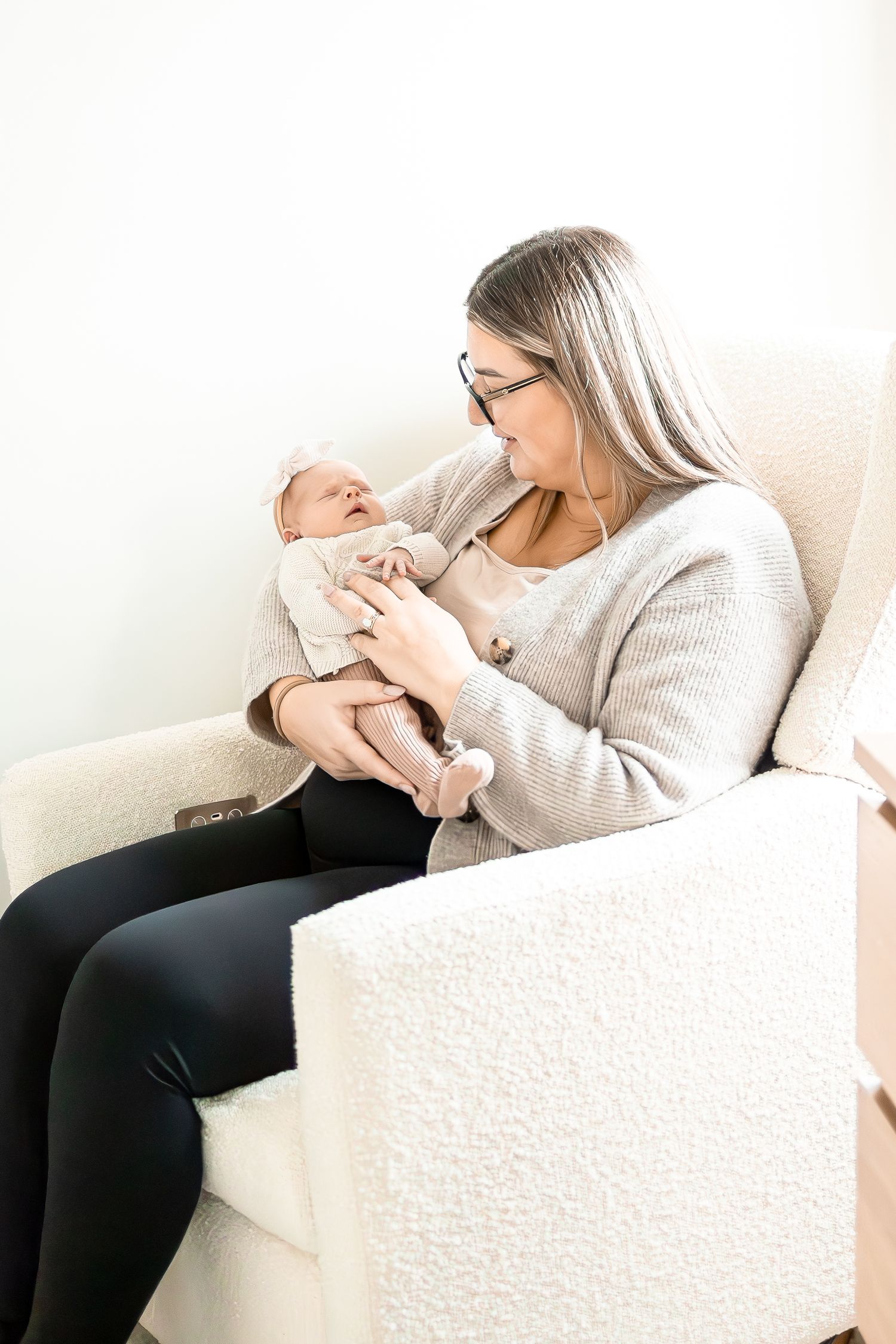 A caregiver in glasses and gray sweater tenderly holds a sleeping newborn in a white outfit.