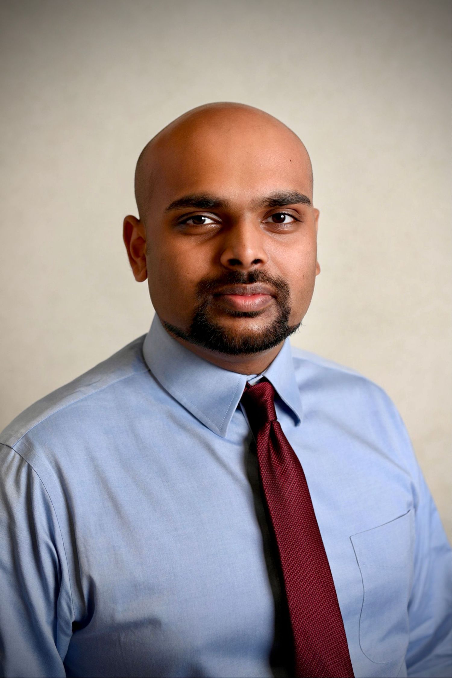 Professional headshot of person in light blue dress shirt and burgundy tie against neutral background.