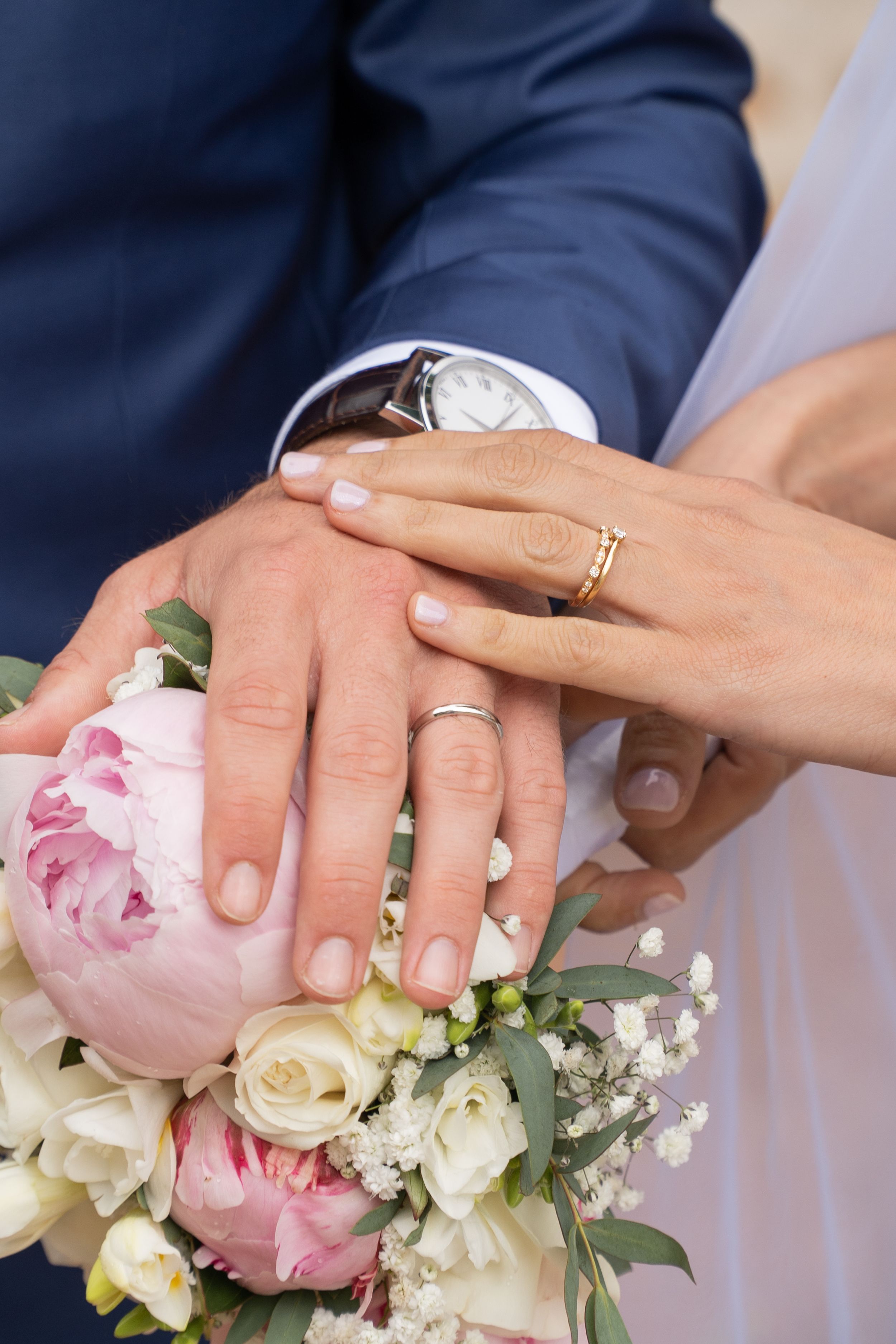 Close-up of wedding rings and hands with pink and white floral bouquet showing romantic wedding details.