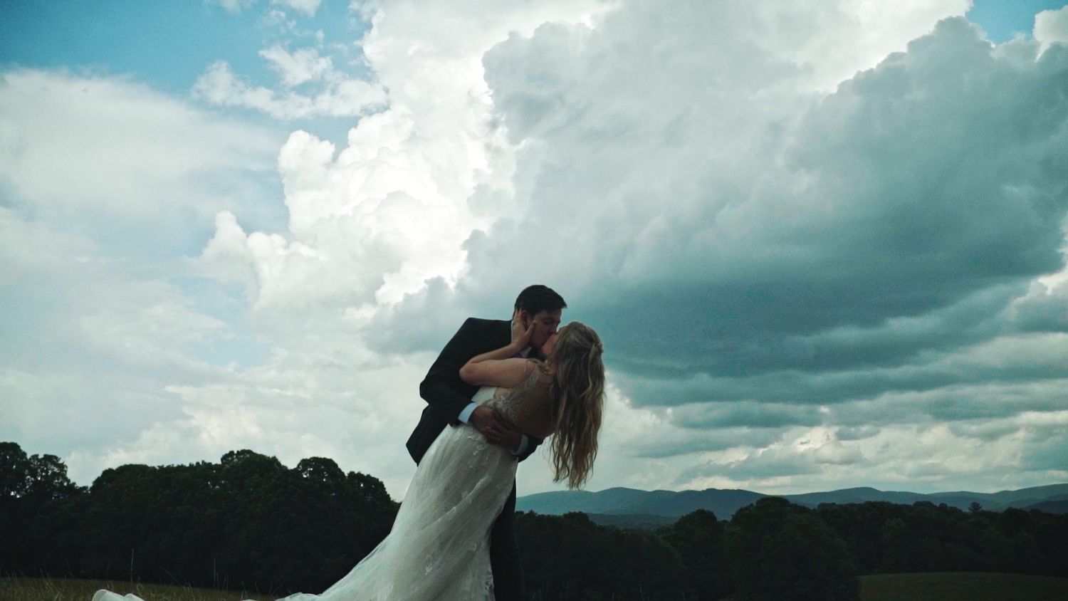 Groom dipping the bride and kissing her with big clouds in the sky