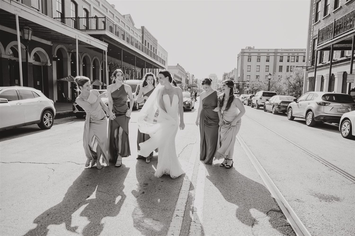 A wedding party walks down a historic street with parked cars on both sides in black and white.