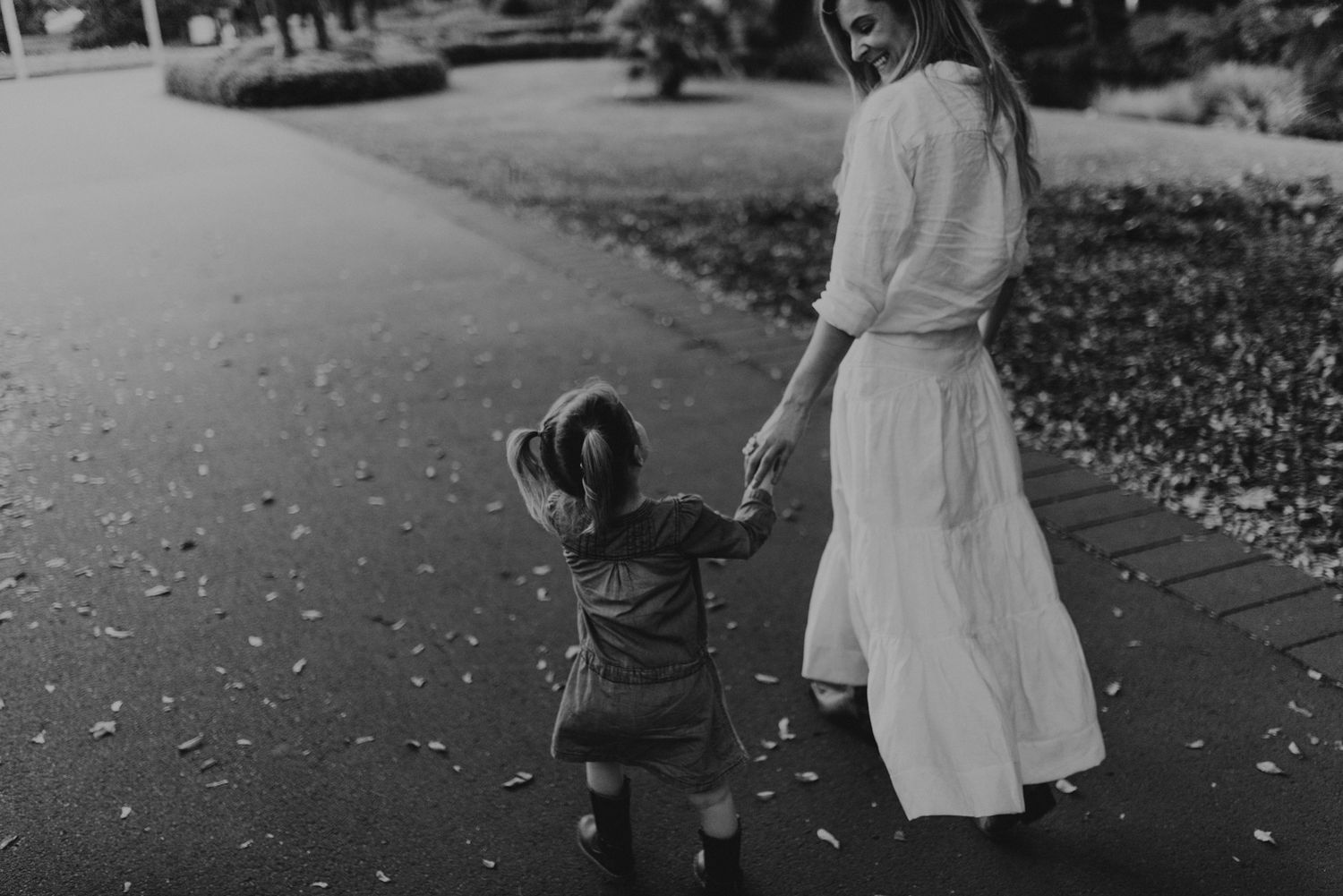 A black and white image shows a tender walking moment between parent and toddler on a leaf-scattered path.