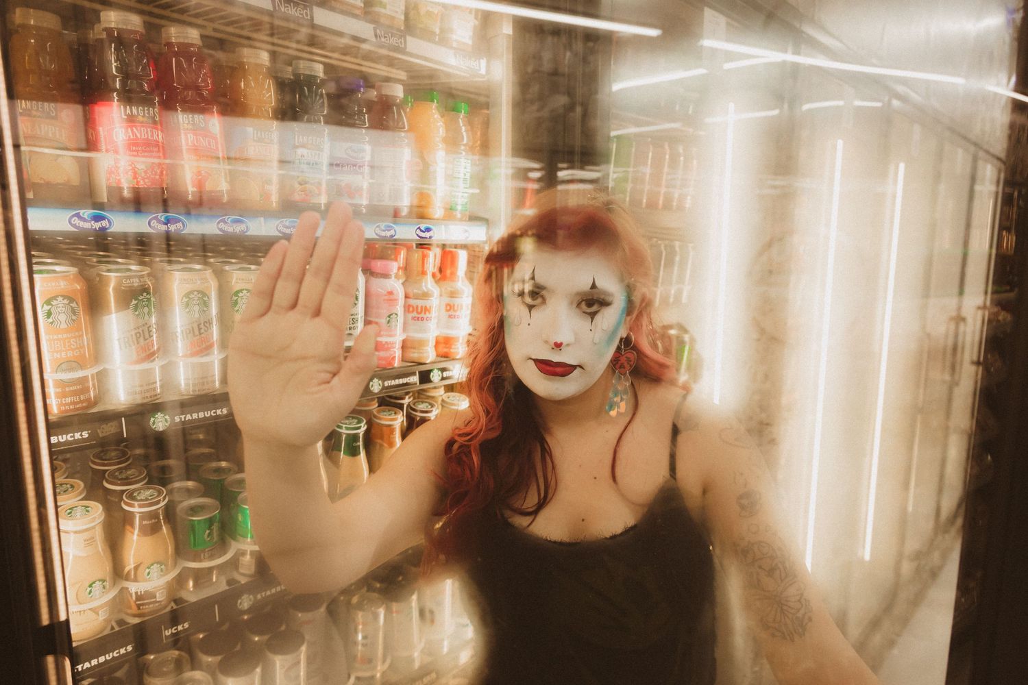 Artistic portrait with dramatic lighting showing a figure with theatrical makeup pressing against glass near a refrigerator display.