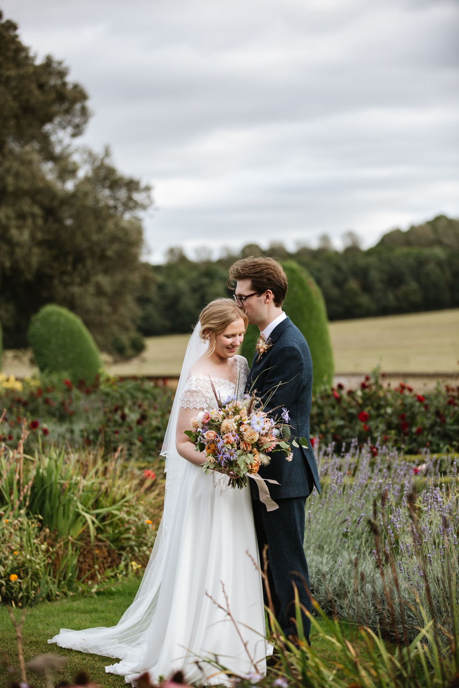 A wedding couple stands together in a lush garden with colorful wildflower bouquet and flowing white dress.