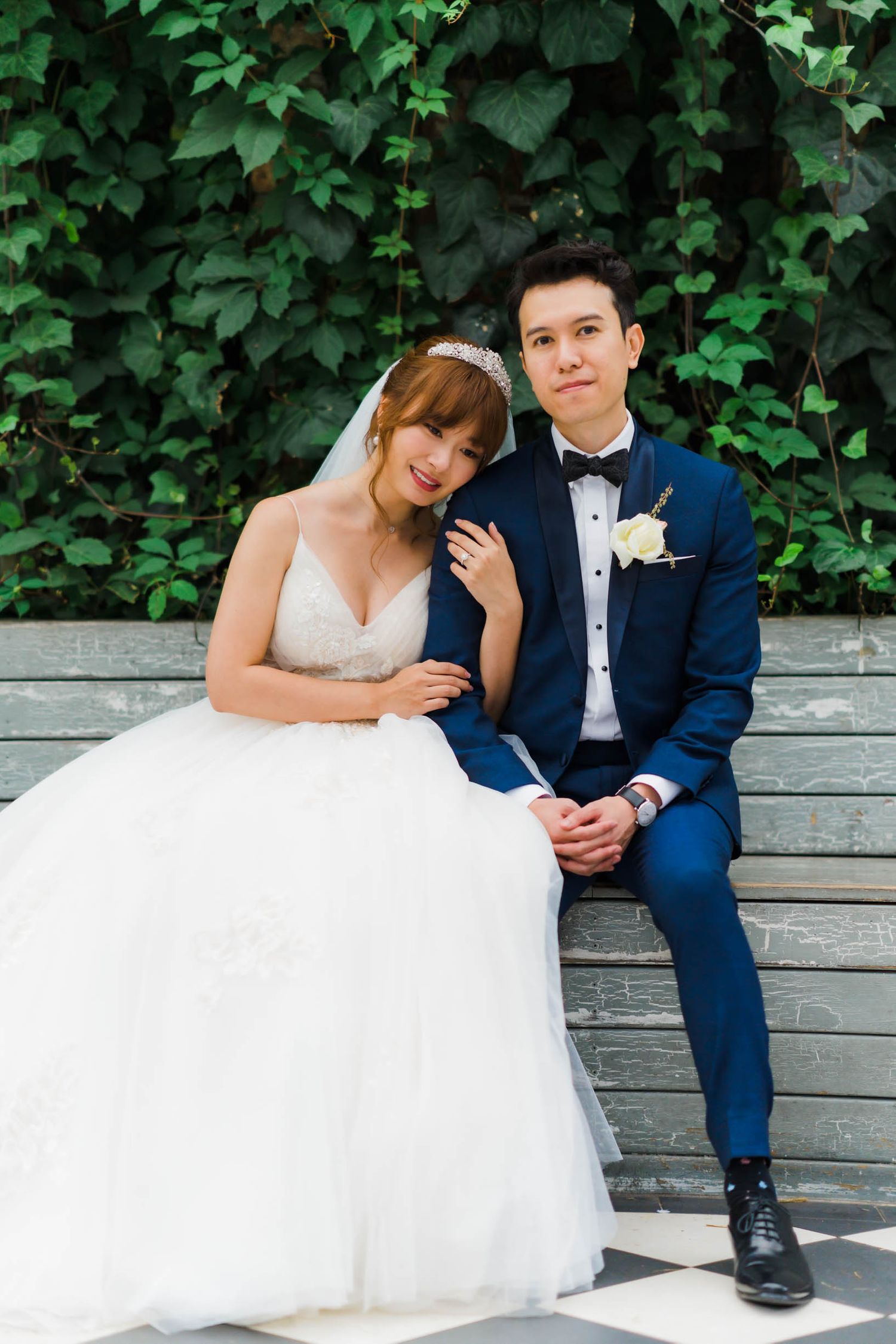 A couple in wedding attire sitting together on wooden steps with ivy-covered wall behind them.